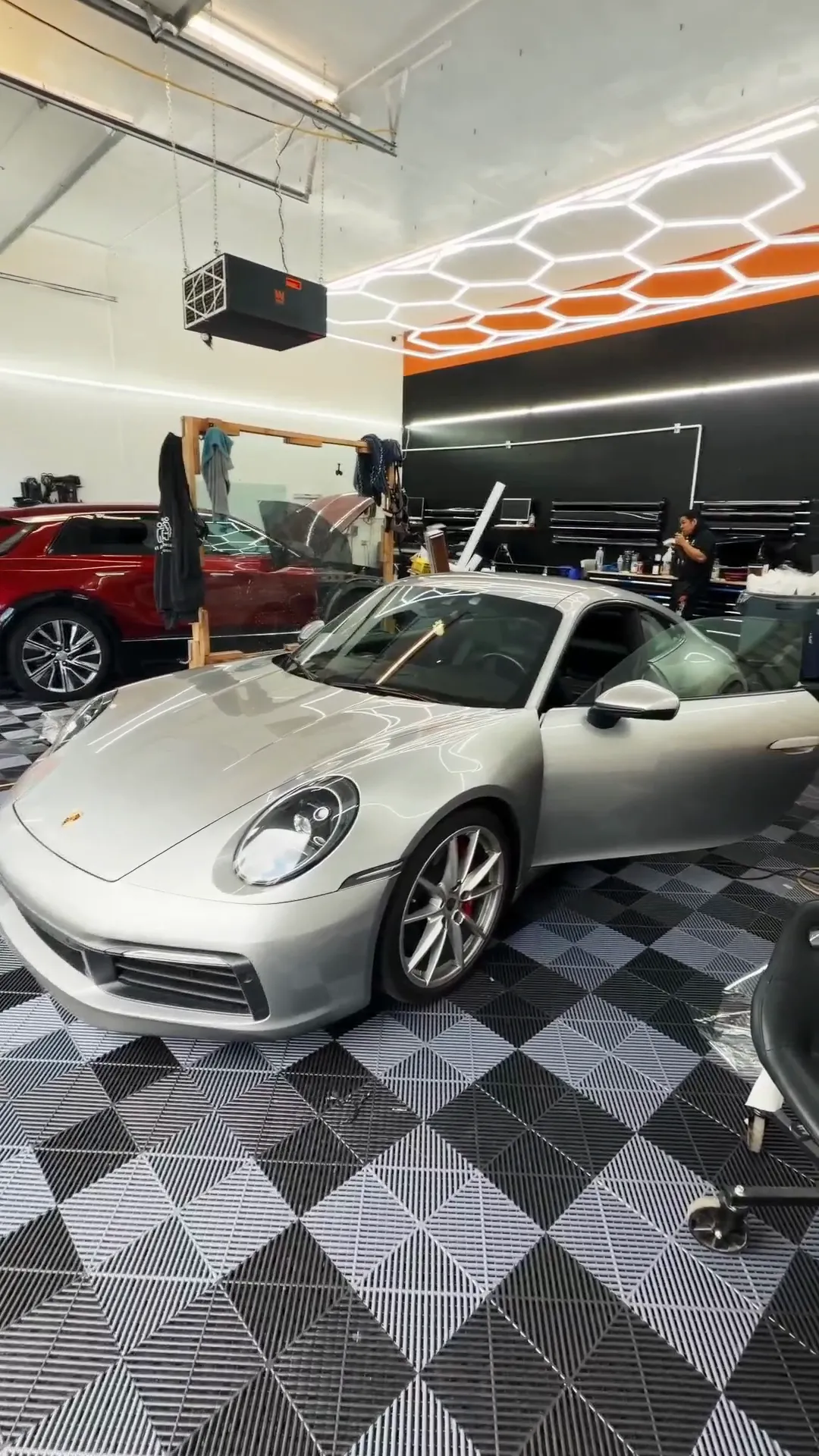 Silver Porsche 911 parked inside A1 Auto Detailing's shop under hexagon ceiling lights, with a technician working in the background