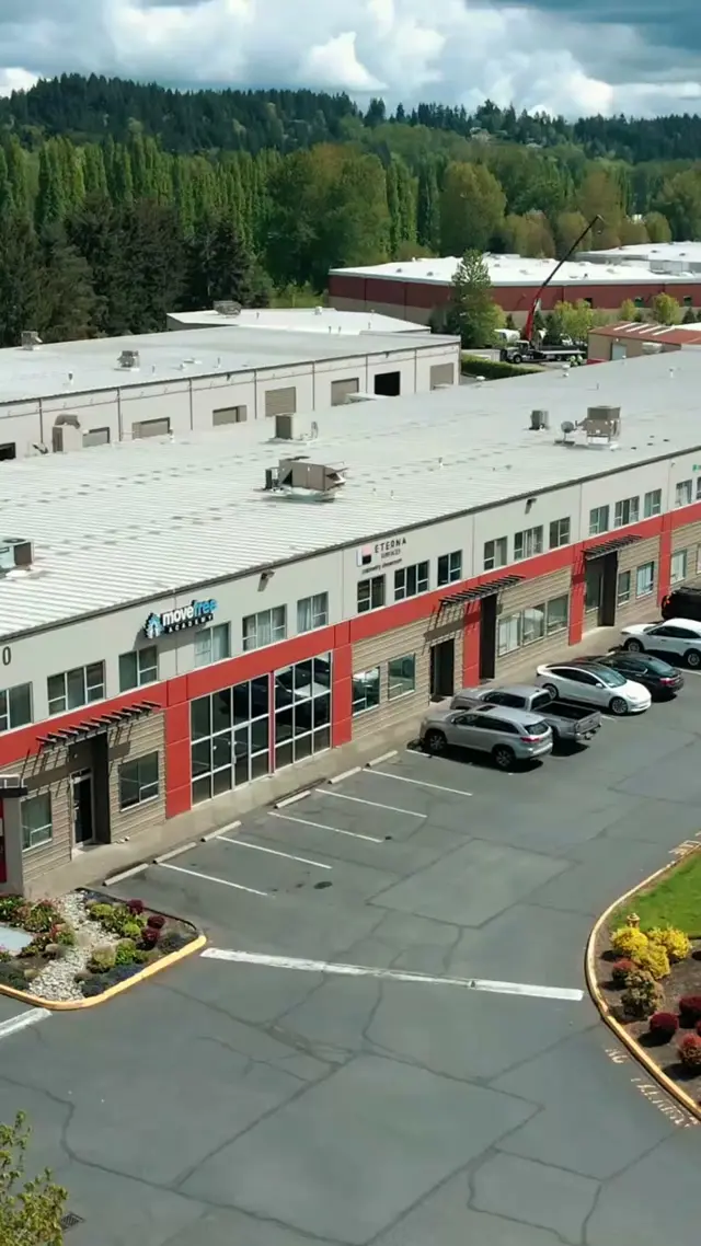 Aerial drone view of the business park building that houses the Eterna Surfaces showroom, a long two-story commercial building with red trim and a parking lot, with evergreen trees behind it