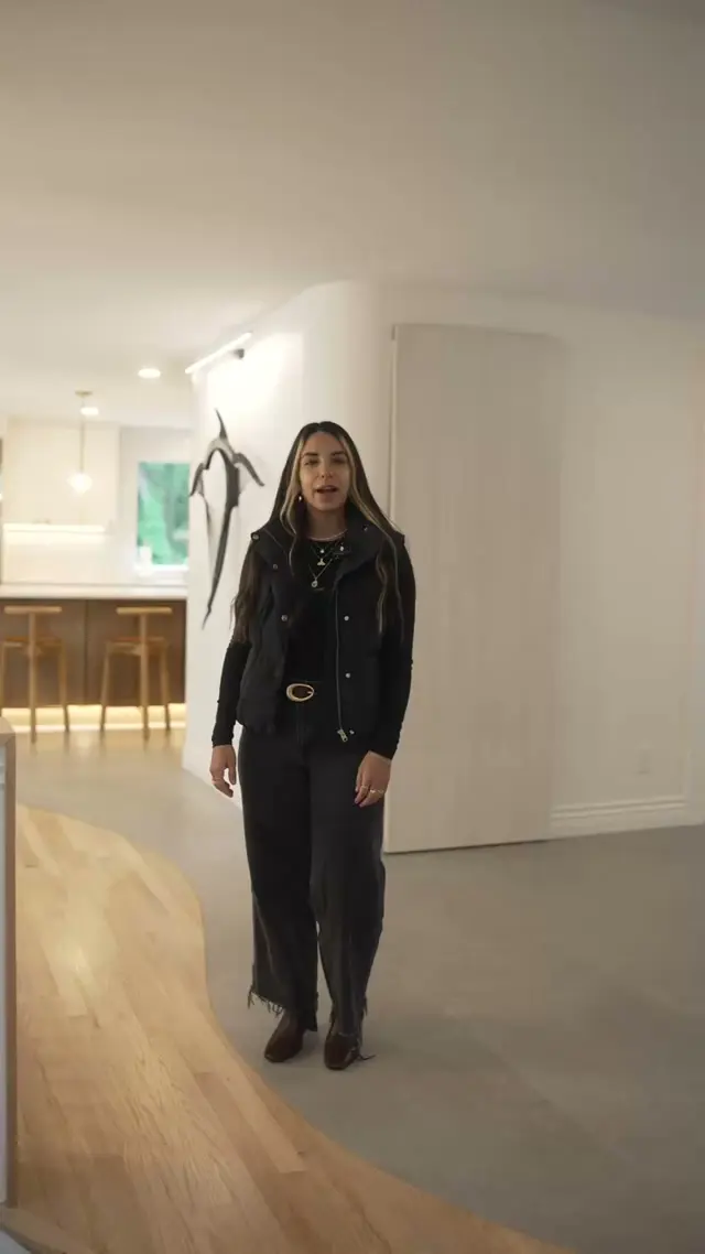 A woman in a black vest talking straight to camera inside a finished Fine Element Construction remodel, with a curved white wall and a kitchen island behind her