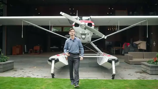 Nazarov Air's owner wearing a lav mic, walking toward the camera in front of a white Piper Super Cub on amphibious floats, parked on a patio in front of a wide open hangar door with furniture inside